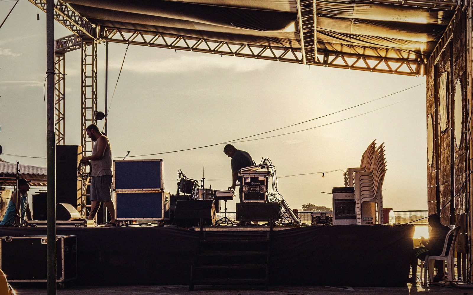 Crew members set up equipment cases and sound gear on an outdoor stage at sunset