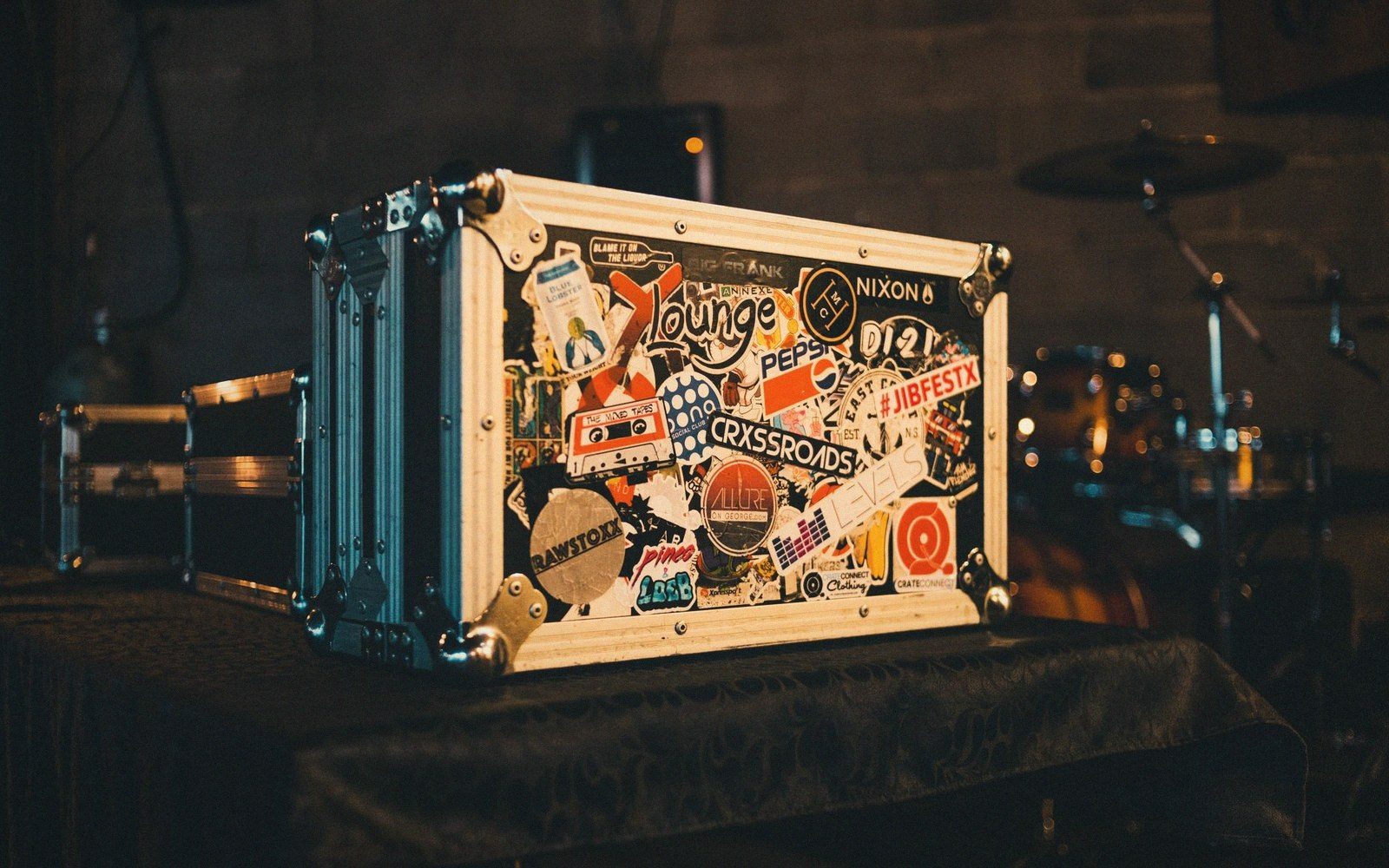 Sticker-covered flight case sits on a stage table near drums and rack equipment