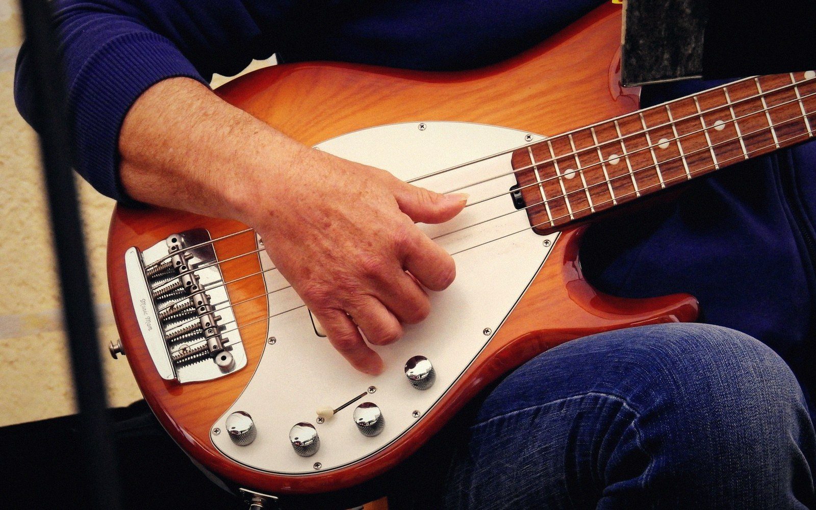 A musician's hand rests on the strings of a sunburst electric bass guitar